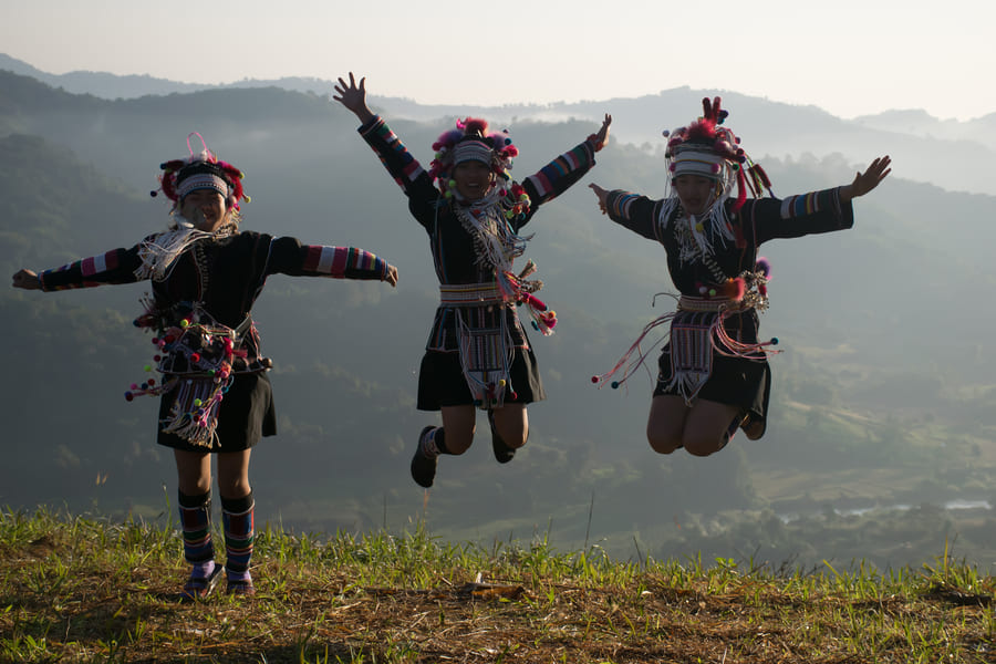 Tres mujeres vestidas con elaborados y coloridos trajes tradicionales de las colinas, con tocados detallados, saltan alegres con los brazos extendidos en la cima de una colina, con un fondo de montañas brumosas y valles bajo una luz suave.