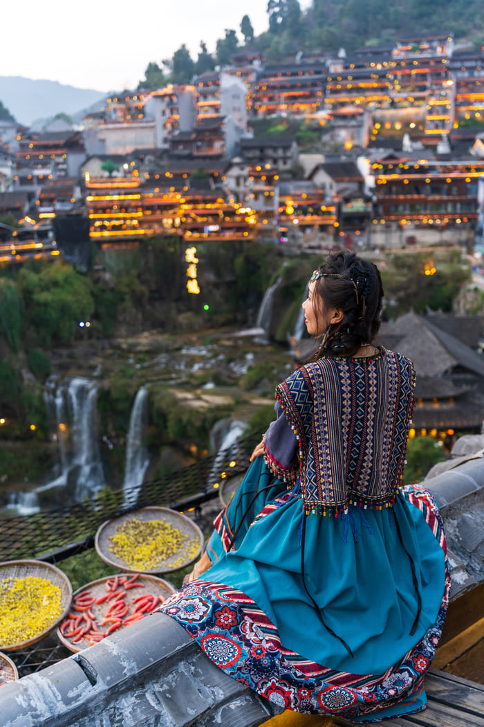 Una mujer recogiendo verduras y mirando un paisaje a lo lejos, de edificios antiguos de China. Vestida de modo tradicional.
