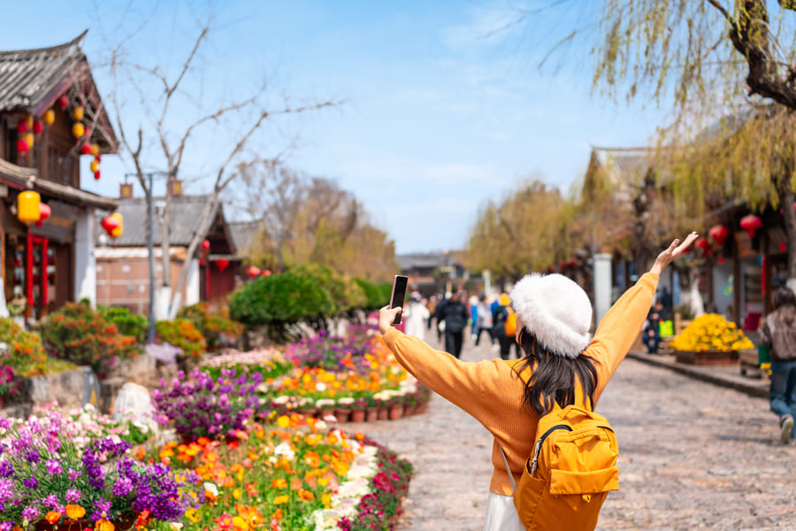 Una persona tomandose fotos sobre un camino, de fondo flores y un edificio antiguo de china en turismo-lijiang