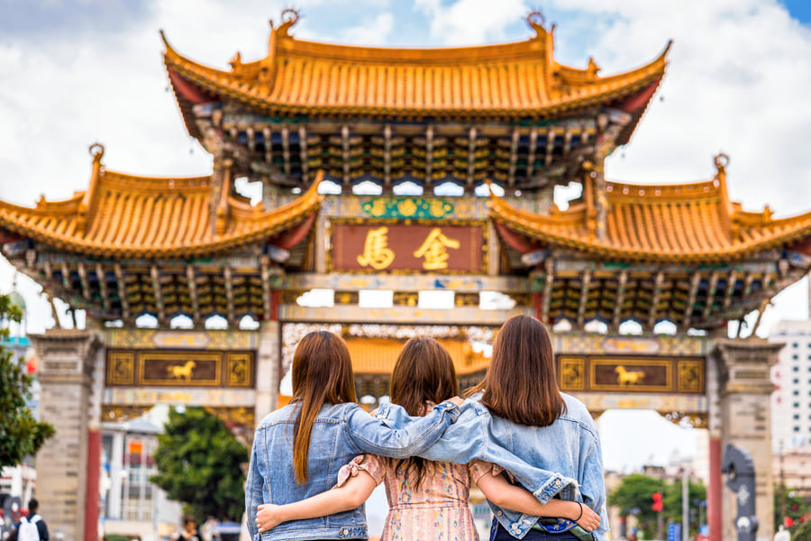 Un viaje de amistad y cultura en Kunming, China. Esta imagen captura a tres amigas compartiendo un momento de contemplación frente al majestuoso e histórico arco ceremonial Jinma, un símbolo de la rica tradición de la ciudad.